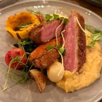 Seared steak with roasted vegetables, polenta, and greens on a white plate.