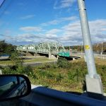 Green truss bridge spans over a highway under a partly cloudy blue sky.