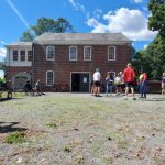 Historic brick building with people gathered on a sunny, grassy field.