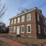 Historic brick building with white trim stands on a grassy lawn under a blue sky.