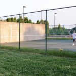 Tennis player serves on outdoor court behind a chain-link fence with backstop.