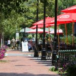 Outdoor cafe scene with red umbrellas, brick walkway, and lush green trees on a sunny day.