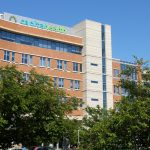 Modern brick hospital building with "Capital Health" signage, partially obscured by green trees.