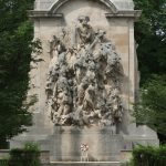 Baroque relief sculpture on a stone monument surrounded by lush green trees.