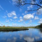 Blue sky with clouds over a calm river flowing through green landscape.