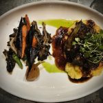 Plated gourmet dish featuring seared meat, roasted vegetables, and bright green sauce.