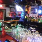 Bar interior with bottles, glassware, and a red counter under dim lighting.