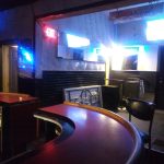Dimly lit bar interior with dark wood counters and neon accents.