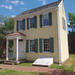 Yellow historic house with green shutters and front porch under a blue sky.