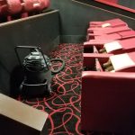 Theater seating rows in a dark auditorium with a black projector on the patterned carpet.