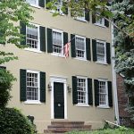 Two-story historic house with green shutters and an American flag displayed.