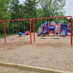 Red swing set and blue playground equipment on a wood chip surface surrounded by trees.
