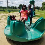 Two children slide down a green water play structure at a sunny park.