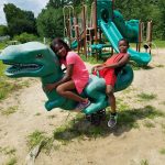 Two children pose on a large, green dinosaur playground structure outdoors.
