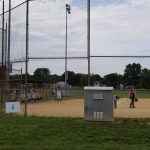 Baseball field with backstop netting, players practicing, and a utility box visible.