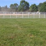 Baseball field with white fencing, green grass foreground, and trees in the background under a blue sky.