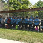 Group of young athletes in uniforms gathered behind a chain-link fence at an outdoor sports field.