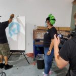 Artist paints on a large white canvas while others watch in an indoor studio setting.