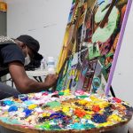 Artist paints a large, colorful abstract canvas in a bright studio setting.