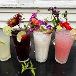 Colorful cocktails with garnishes on a black table against a white wooden background.