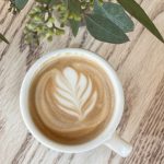 Latte art in a white mug on light wood, with green foliage overhead.
