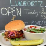 Burger and salad served with a pickle on a plate, in front of a chalkboard menu.
