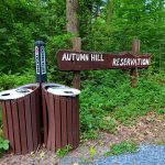 Recycling and trash bins near a wooden sign marking Autumn Hill Reservation in a wooded area.