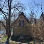 Old stone cabin by a pond, surrounded by bare trees and dry brush.