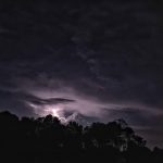 Dark, stormy night sky with a bright flash of lightning illuminating heavy clouds over silhouetted trees.