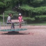 Man and child on a merry-go-round in a park setting with trees in the background.