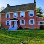 Red colonial house with white trim and shutters on a sunny, grassy lawn.