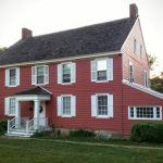 Red clapboard historic house with white trim and dark roof, set on a green lawn.