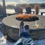 Person's legs resting near a fire pit outdoors in the snow during winter.