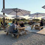 People gather under striped umbrellas at an outdoor event with picnic tables.
