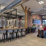 Brewery interior with stainless steel tanks, bar area, and patrons enjoying drinks.
