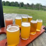 Assortment of craft beers in glasses displayed on a wooden serving tray outdoors.