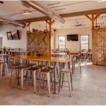 Rustic industrial dining hall with long wooden tables, high ceilings, and exposed beams.