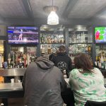 People watching sports on TVs in a dimly lit bar setting with shelves of liquor.