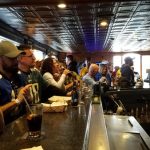 Crowded bar interior with patrons socializing at the counter under a patterned ceiling.