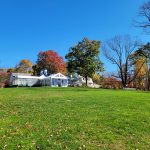 Historic white farmhouse nestled among colorful autumn trees under a bright blue sky.