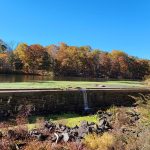 Small waterfall flows over a stone dam into a calm, tree-lined pond under a blue sky.