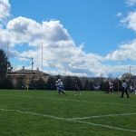 Football game in progress on a sunny, grassy field under a partly cloudy blue sky.