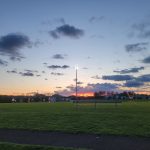 Dusk over a grassy sports field with stadium lights on and colorful sunset clouds.