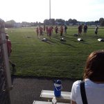 Soccer practice on a sunny field with players and stadium lights visible.