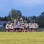Lacrosse players line up on a grassy field with sticks, under a blue sky.