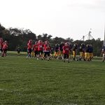 Football players in uniforms run across a green field during an outdoor game.