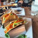 Two toasted sandwiches with avocado and toppings served on a white plate at a restaurant.