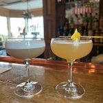 Two cocktails in coupe glasses on a wooden bar, with a well-stocked liquor shelf in the background.