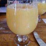 Two glasses of yellow beverage on a wooden table, with a person blurred in the background.