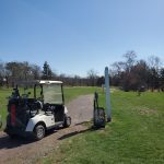 Golf cart parked on a paved path in a sunny, grassy golf course setting.